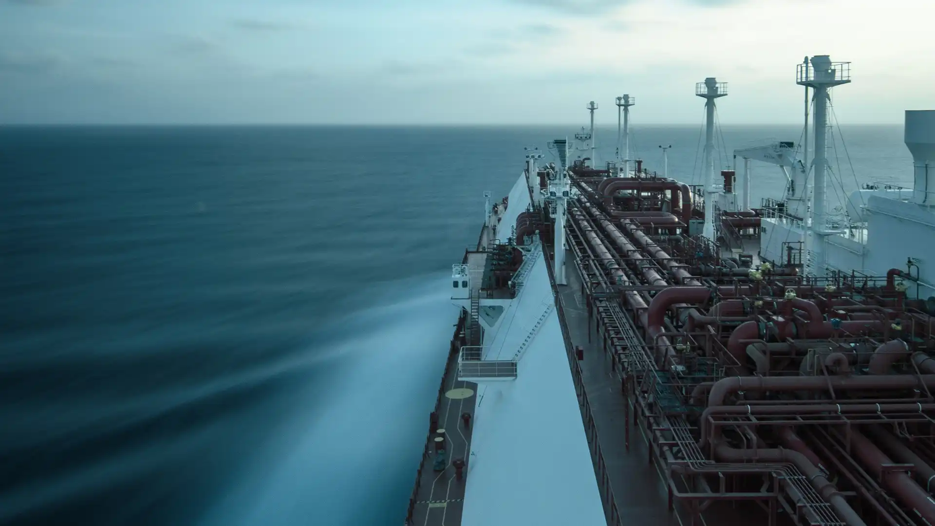 Long-exposure view from an industrial tanker ship at sea, showing pipelines, calm ocean waters and a misty blue horizon at dusk.